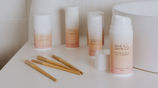 Skincare products from "She & I Skin Co." are arranged on a white surface. The bottles feature gradient peach-to-white labels. Several makeup brushes with light bristles and gold handles lie nearby.