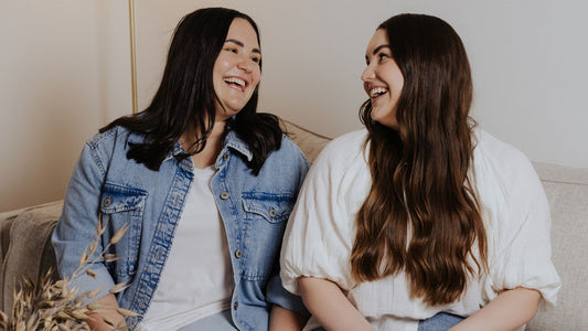 The owners of She & I Skin Co. are sitting on a couch, smiling and looking at each other. One wears a denim jacket over a white shirt, and the other wears a white blouse