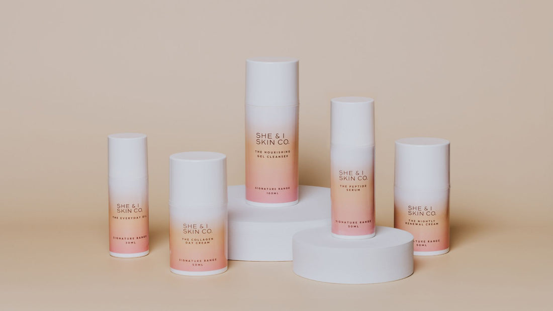 Six skincare products from "She & I Skin Co." are displayed on a beige background. The bottles feature gradient peach-to-white labels and are arranged neatly on white circular platforms.