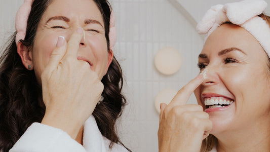 Two women wearing soft pink headbands playfully apply cream to each other’s noses. Both are smiling and dressed in white robes, enjoying a lighthearted skincare moment.