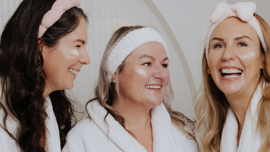 Three women wearing white robes and headbands smile together with skincare cream applied under their eyes. They appear relaxed, enjoying a shared beauty or self-care moment.