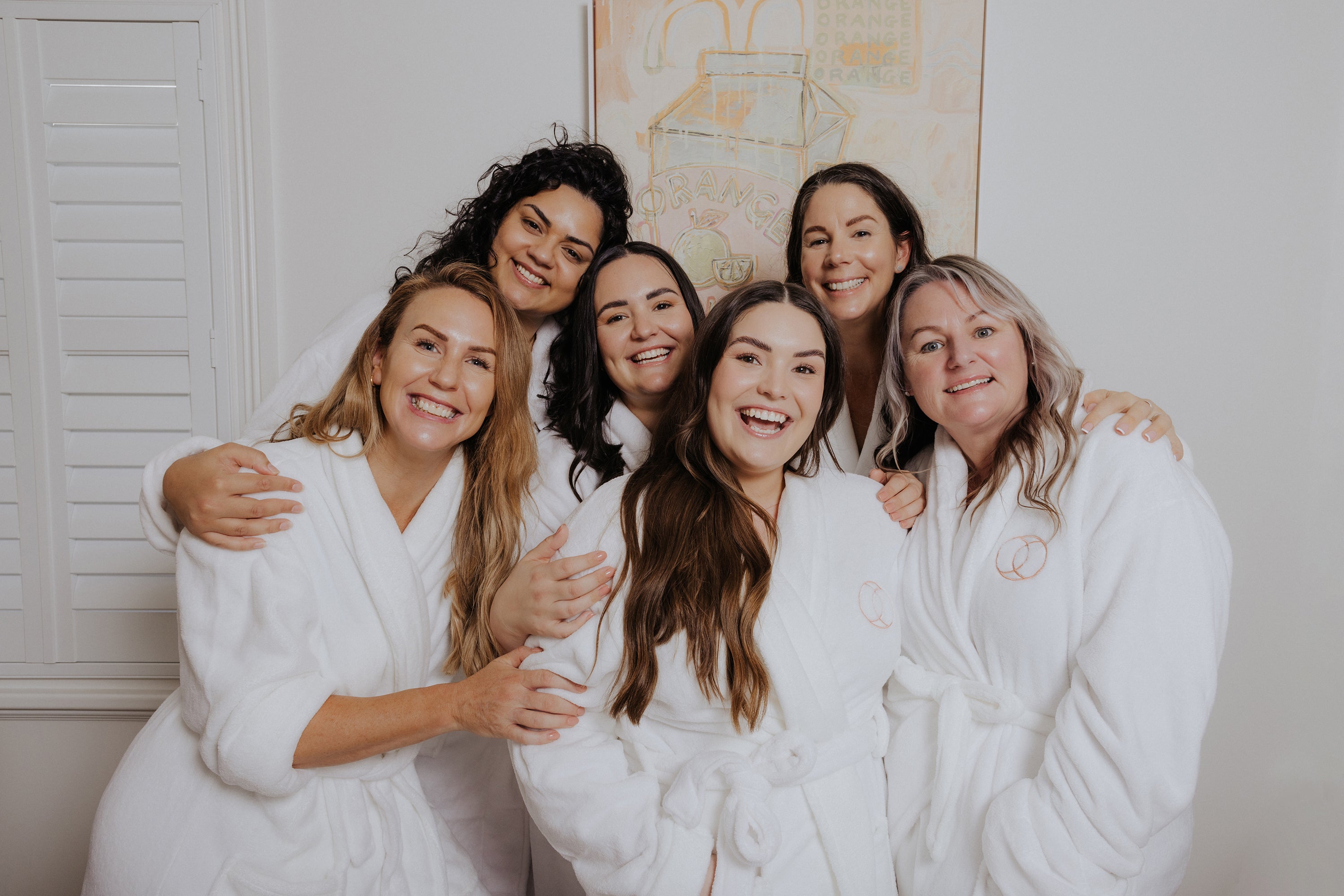 A group of six smiling women in white robes stand together in a bright room, radiating joy and warmth, with soft art and shutters in the background.