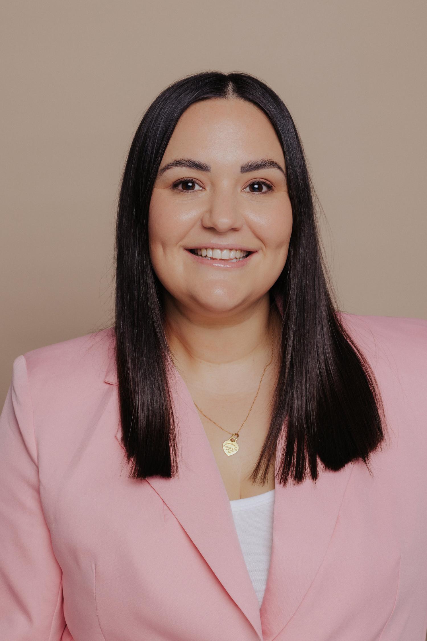 A woman, Sarah Slatter, with straight dark brown hair smiles warmly, wearing a pink blazer and a gold heart-shaped pendant necklace against a beige background.