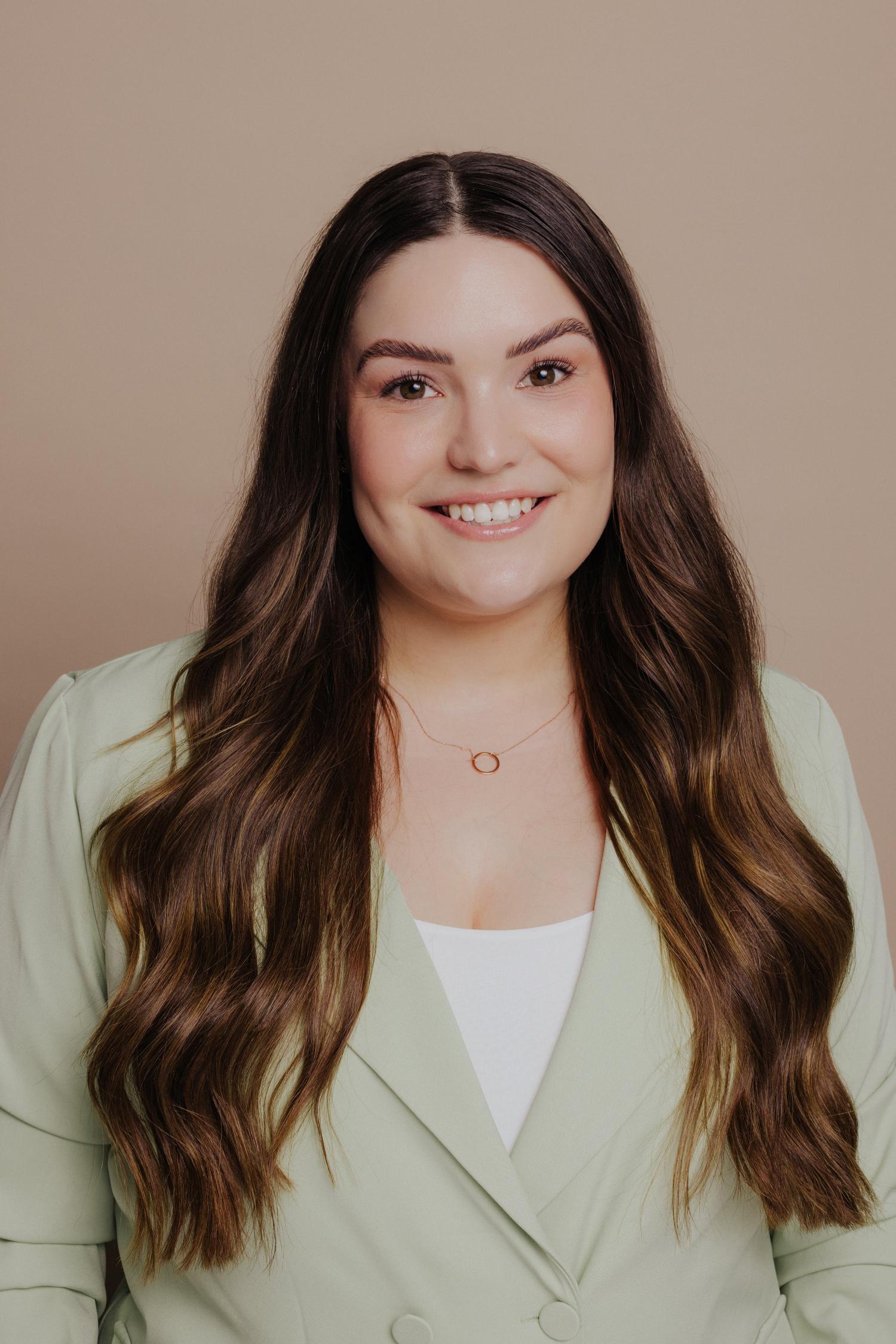 A woman, Ella Slatter, with long, wavy brown hair smiles warmly, wearing a light green blazer and a simple gold circle necklace against a beige background.