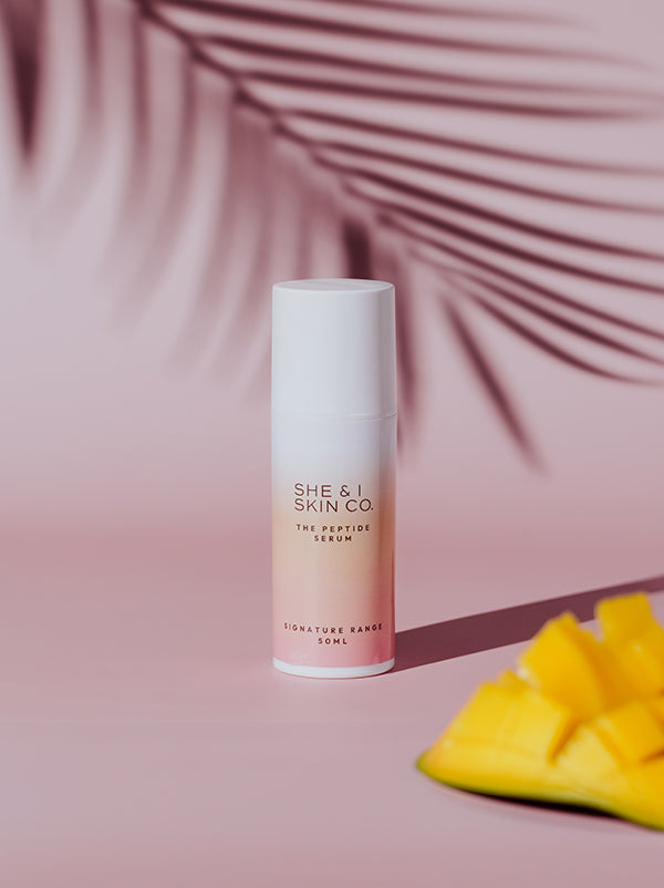 She & I Skin Co. "The Peptide Serum," 50ml, displayed alongside fresh mango slices and tropical palm shadows. A refreshing and luxurious addition to any skincare routine.